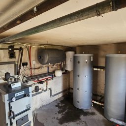 Tetiana Senkiv - Piwnica z zainstalowanym piecem na paliwo stałe, dwoma szarymi zbiornikami akumulacyjnymi i rurami. Widoczne ślady wilgoci na podłodze.