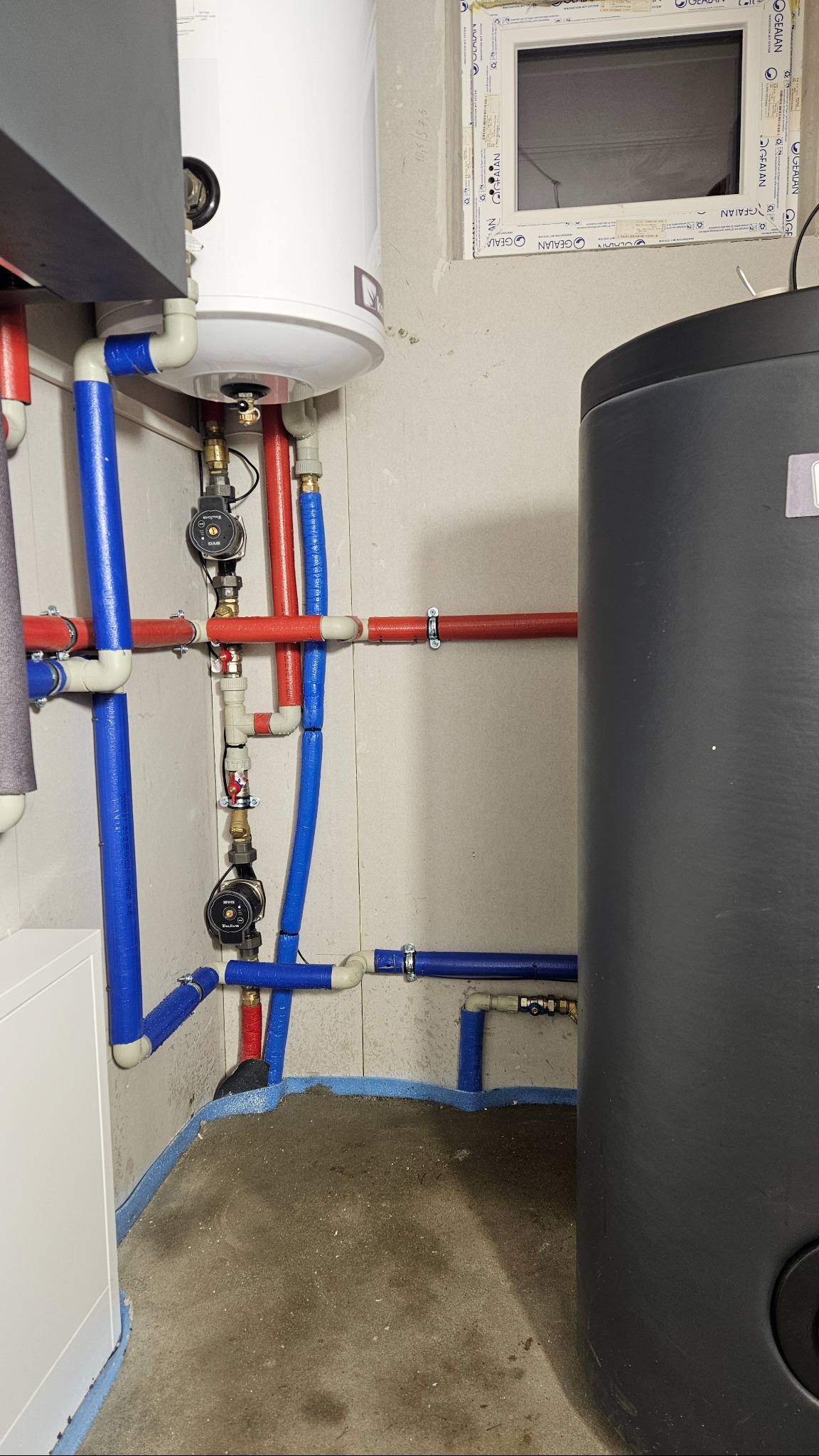 Złożona instalacja grzewcza z rurami w izolacji (czerwone i niebieskie) oraz zbiornikiem akumulacyjnym. Widoczne pompy obiegowe i fragment okna w tle.