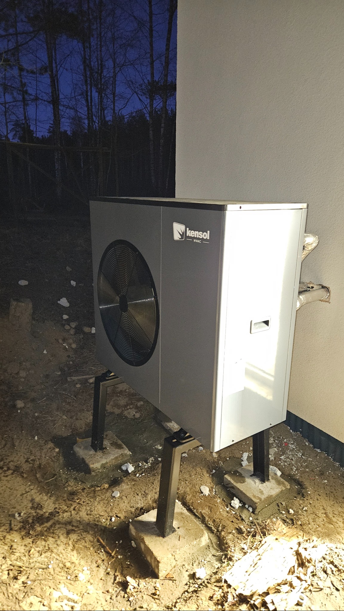 Szara pompa ciepła Kensol HVAC zamontowana na zewnątrz budynku, widoczne rury przyłączeniowe i betonowe podstawy. W tle las i wieczorne niebo.