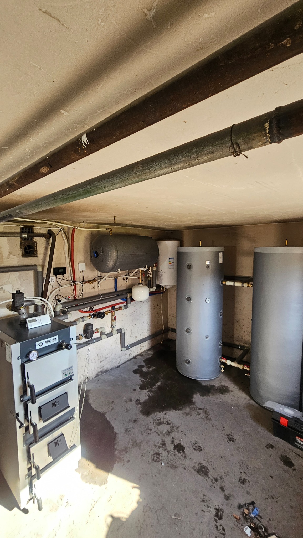 Piwnica z zainstalowanym piecem na paliwo stałe, dwoma szarymi zbiornikami akumulacyjnymi i rurami. Widoczne ślady wilgoci na podłodze.