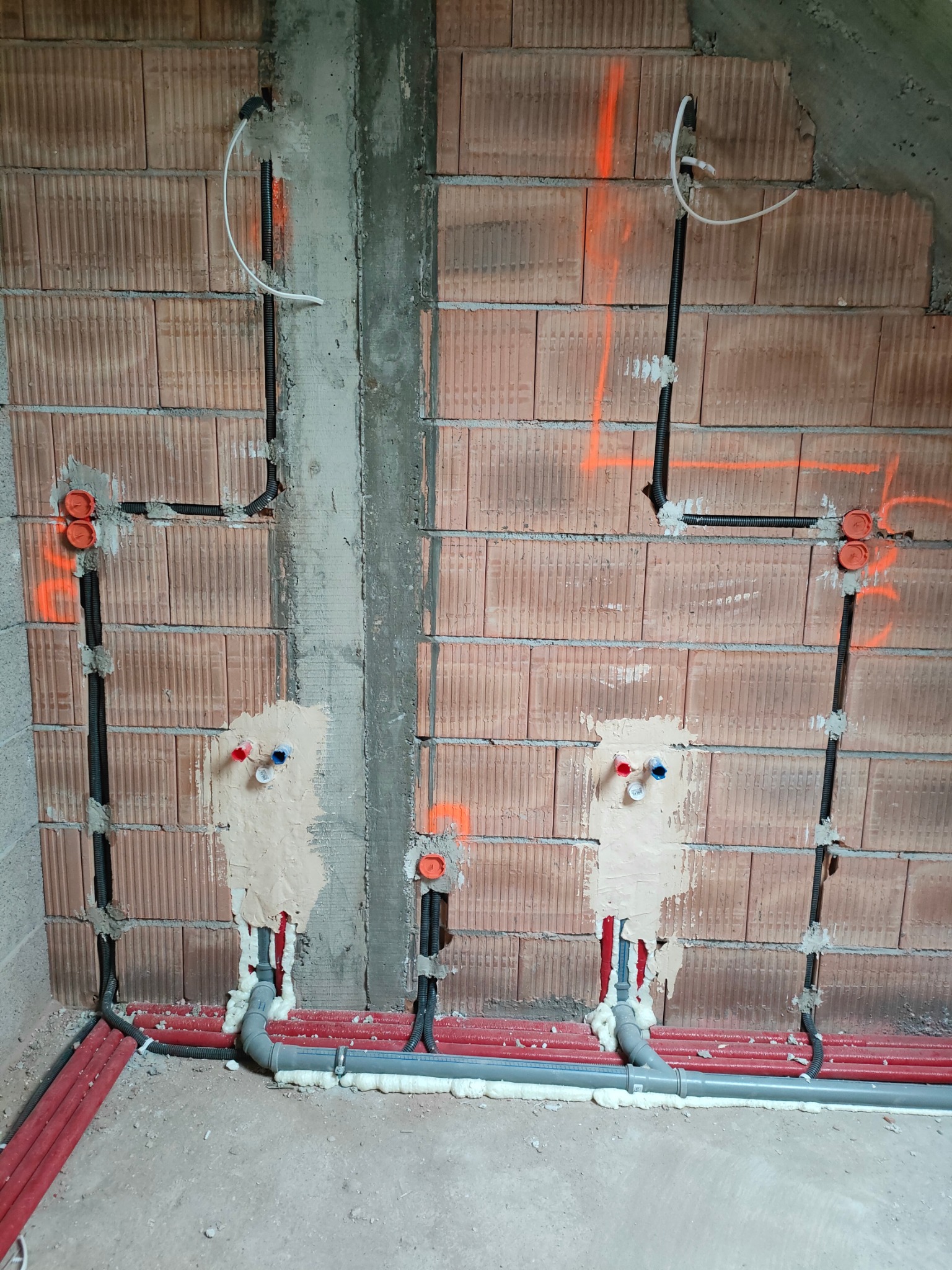 Ściana z instalacją elektryczną i hydrauliczną w trakcie montażu. Widoczne kable w peszlach, rury czerwone i szare, puszki elektryczne oraz pianka montażowa.