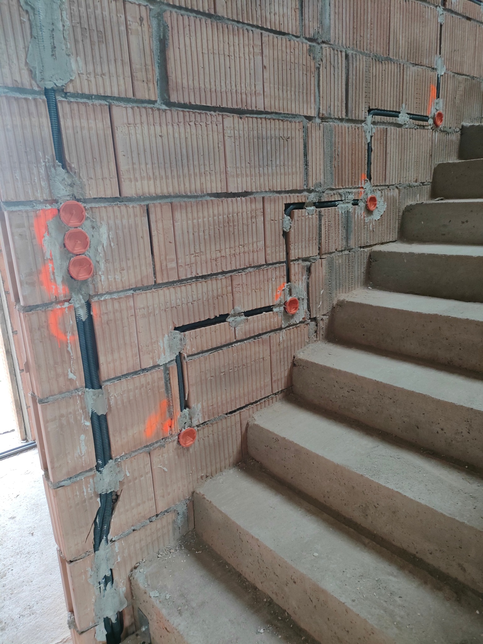 Czarne rury instalacji elektrycznej w bruzdach ceglanej ściany, z pomarańczowymi puszkami pod gniazdka, widoczne betonowe schody w surowym stanie.