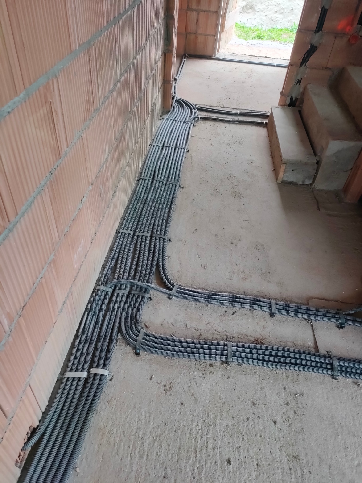 Szara, karbowana rura instalacyjna ułożona wzdłuż ceglanej ściany w surowym wnętrzu. Rury przymocowane są do ściany za pomocą białych uchwytów.