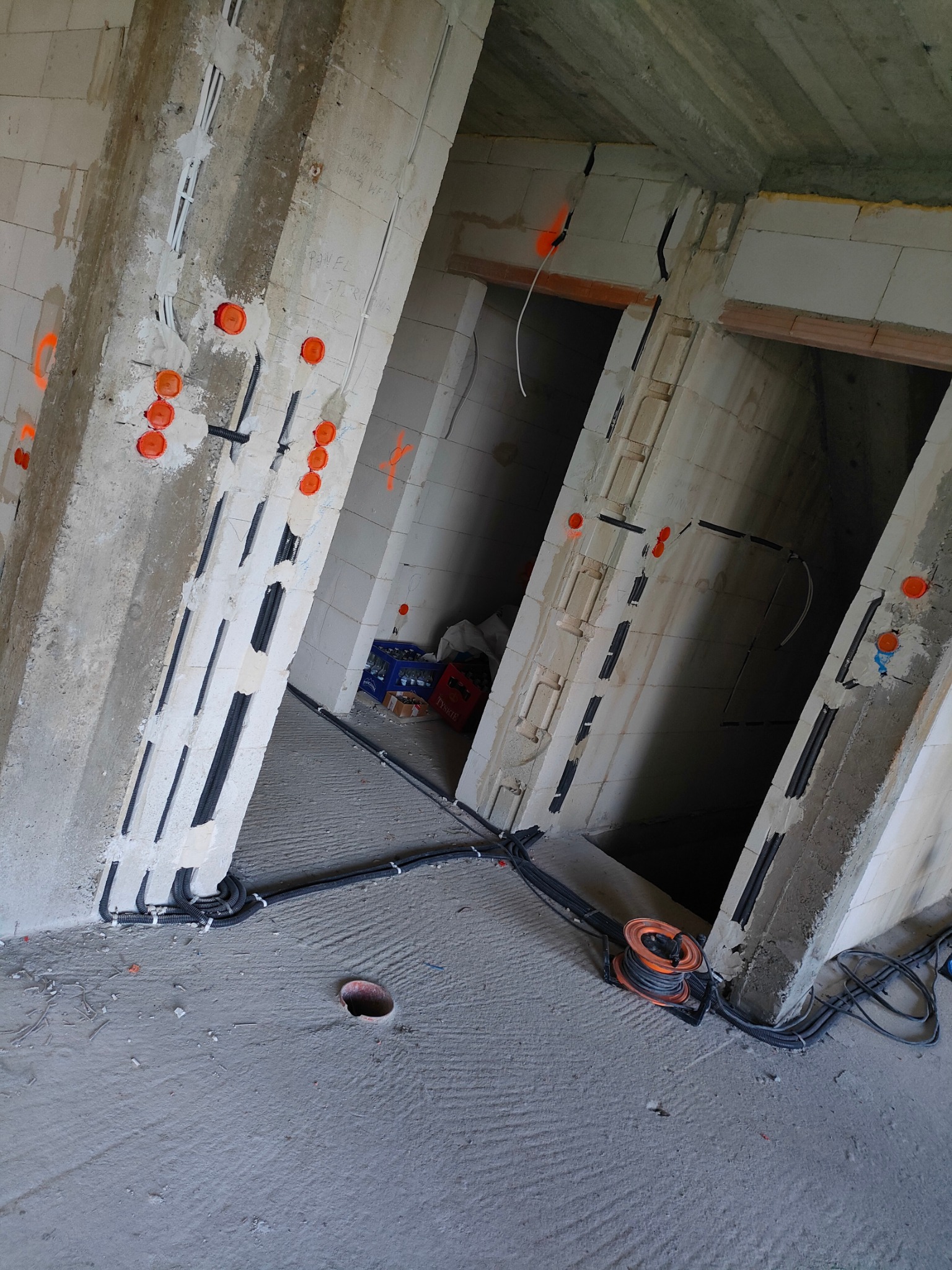 Surowy stan: instalacja elektryczna w bruzdach ścian z betonu komórkowego, pomarańczowe puszki, kable w peszlach, zwój kabla na podłodze. Widok z góry.