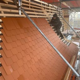 KATPOL - Montaż brązowej dachówki karpiówki na drewnianej konstrukcji dachu, widok z rusztowania. Dekarz w trakcie pracy, w tle zabudowa miejska.