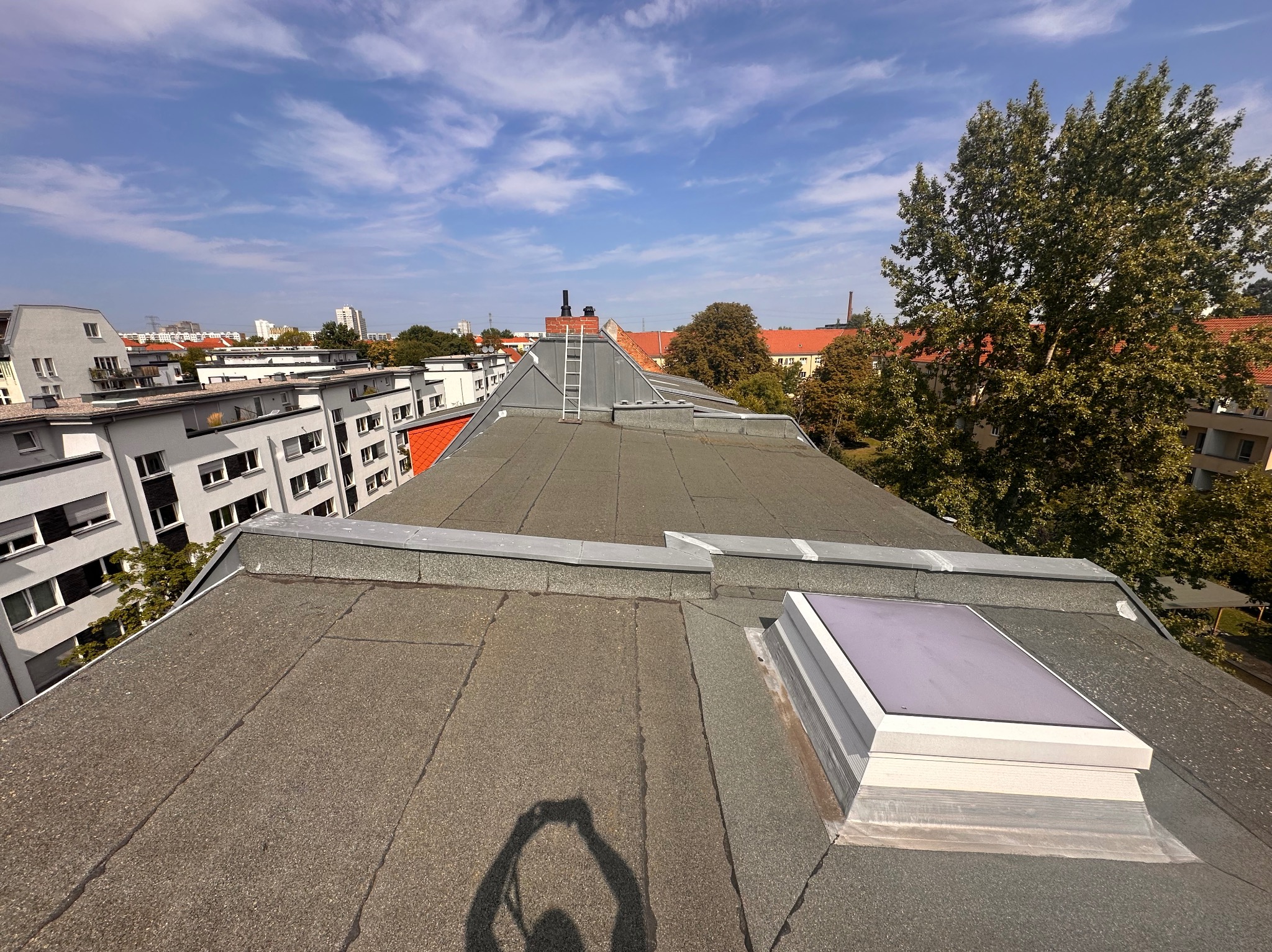 Widok z góry na płaski dach pokryty papą, z widocznym świetlikiem i kominem z drabiną. W tle zabudowa miejska i błękitne niebo z chmurami.