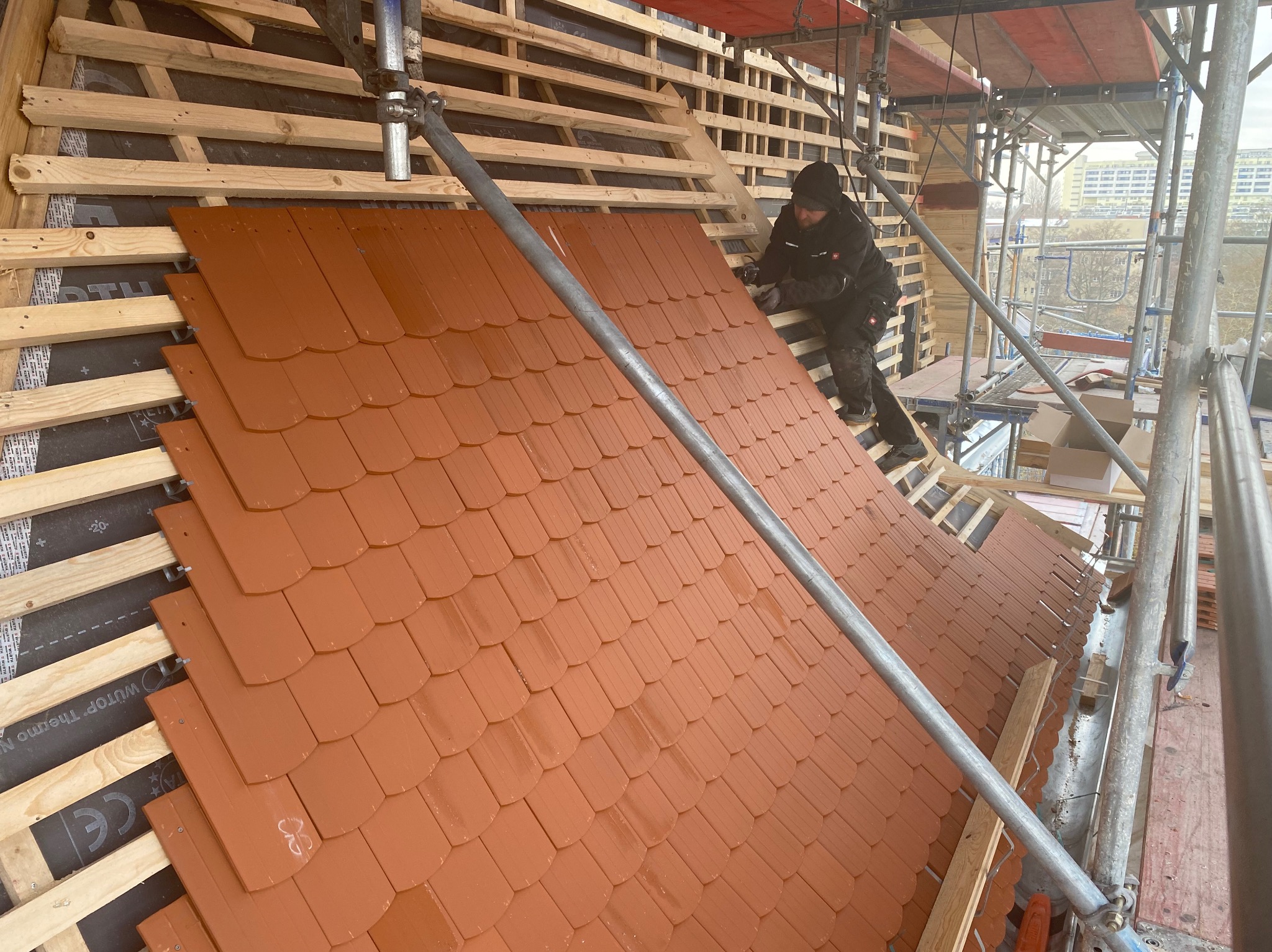 Montaż brązowej dachówki karpiówki na drewnianej konstrukcji dachu, widok z rusztowania. Dekarz w trakcie pracy, w tle zabudowa miejska.