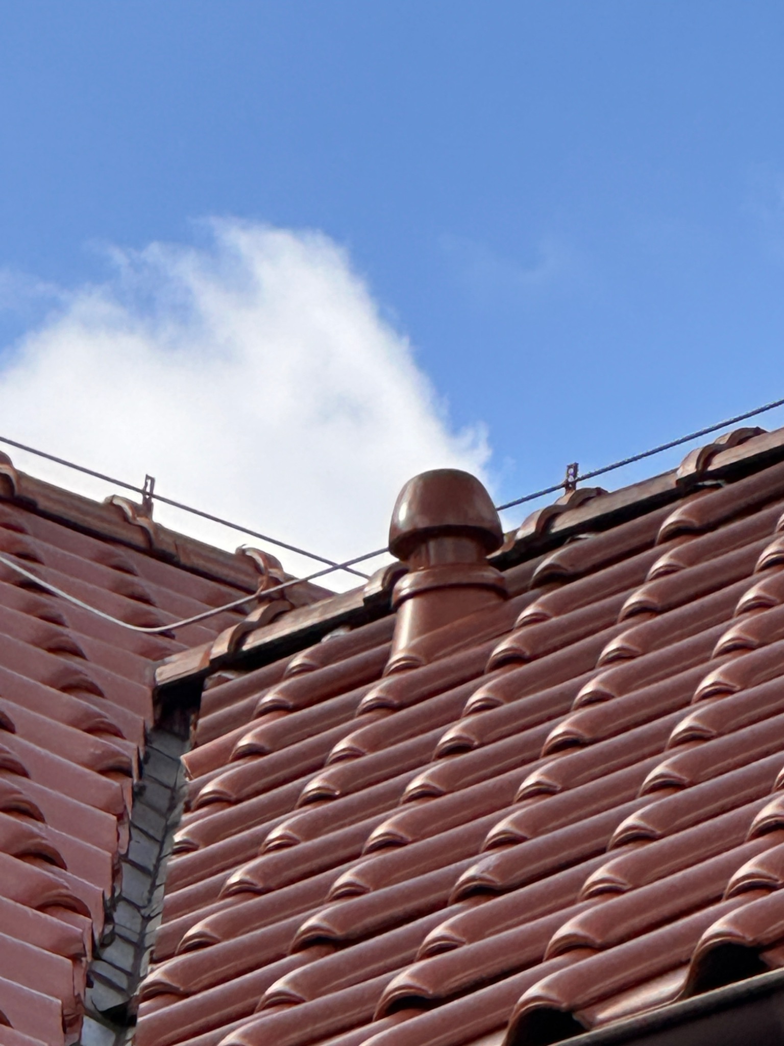 Czerwona dachówka ceramiczna z odpowietrznikiem na tle błękitnego nieba z chmurami. Widoczne zabezpieczenie przeciwśniegowe.