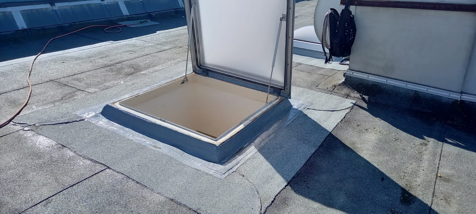 Otwarty świetlik dachowy na płaskim dachu pokrytym papą. Widoczne uszczelnienia i obróbki blacharskie wokół świetlika. W tle widoczny element instalacji i plecak.