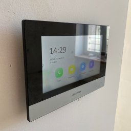 AVASTO Marceli Wikliński - Zamontowany wideodomofon Hikvision na białej ścianie, widoczny ekran z datą i godziną, ikony połączeń i ustawień. Minimalistyczny design, nowoczesny wygląd.