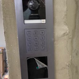 AVASTO Marceli Wikliński - Szary panel domofonu Hikvision z klawiaturą numeryczną i kamerą, zamontowany na surowej ścianie. Częściowo zdjęta folia ochronna na dolnym panelu.