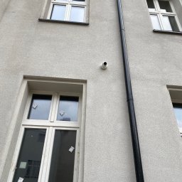 AVASTO Marceli Wikliński - Kamera monitoringu na elewacji budynku z oknami i rurą spustową. Ujęcie z dołu, widoczne okna na różnych kondygnacjach. Szara elewacja.