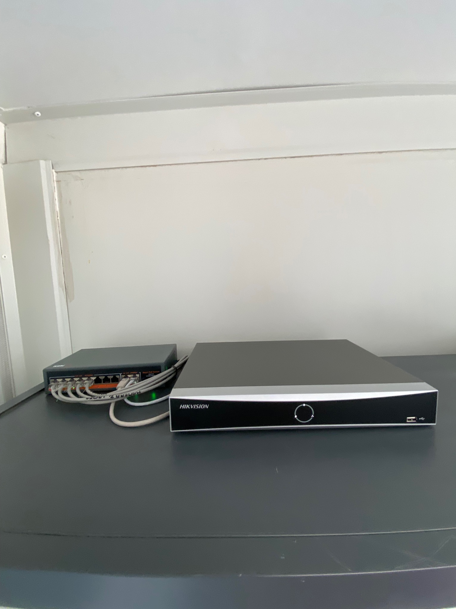 Zamontowany rejestrator Hikvision obok switcha z podłączonymi kablami sieciowymi, na tle białej ściany. Element systemu monitoringu.