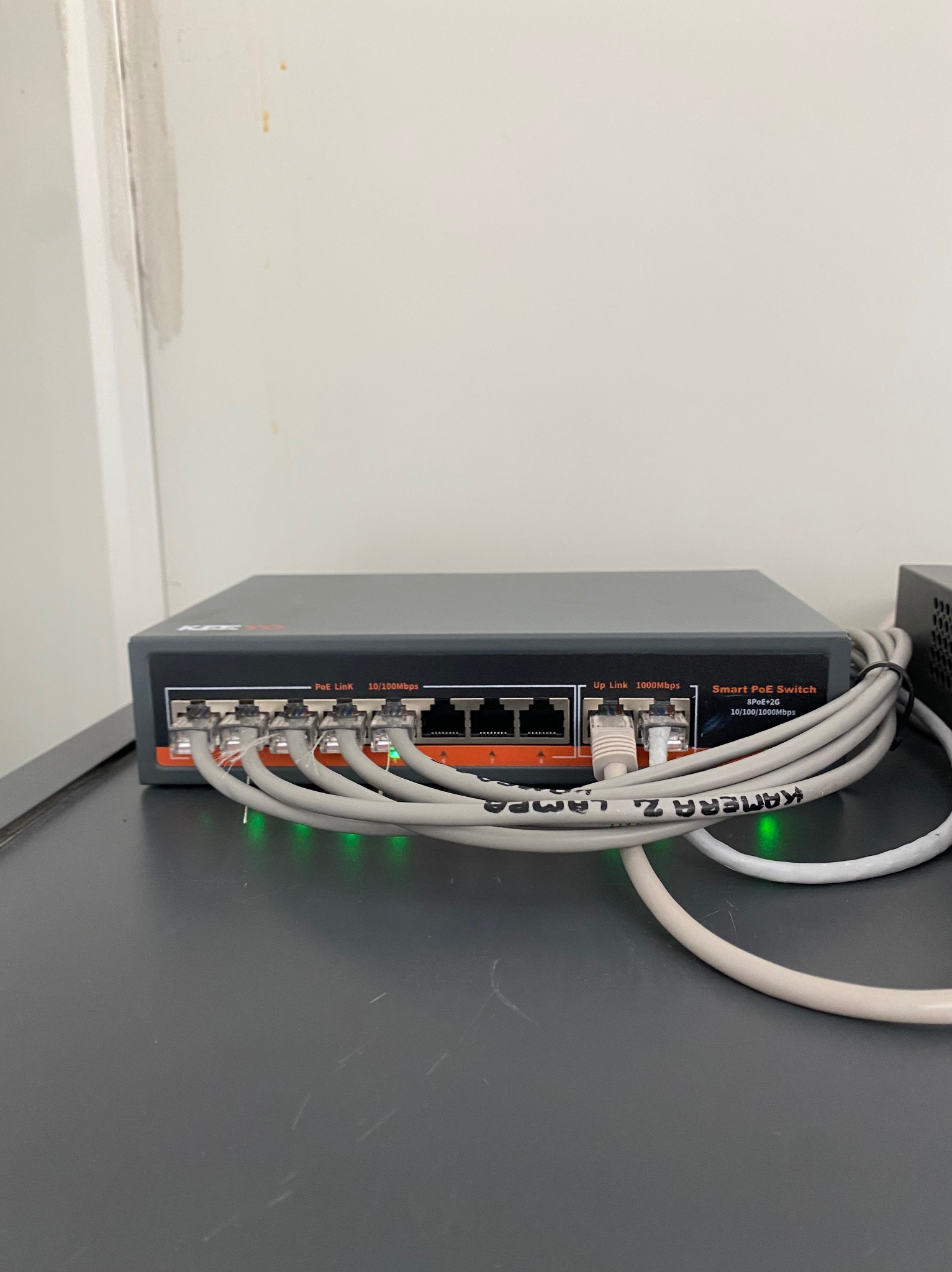 Smart PoE Switch KPC z podłączonymi kablami Ethernet, diody sygnalizacyjne świecą. Kabel oznaczony 'KAMERA Z. LAMPA'. Widok z bliska na urządzenie.