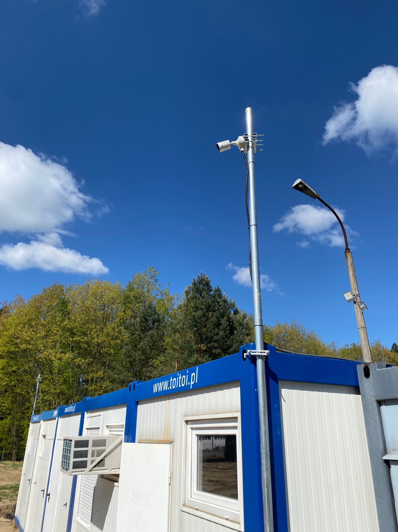 Kamera monitoringu na słupie przy kontenerze biurowym z klimatyzacją na tle błękitnego nieba i drzew. Widoczny adres strony internetowej na kontenerze.