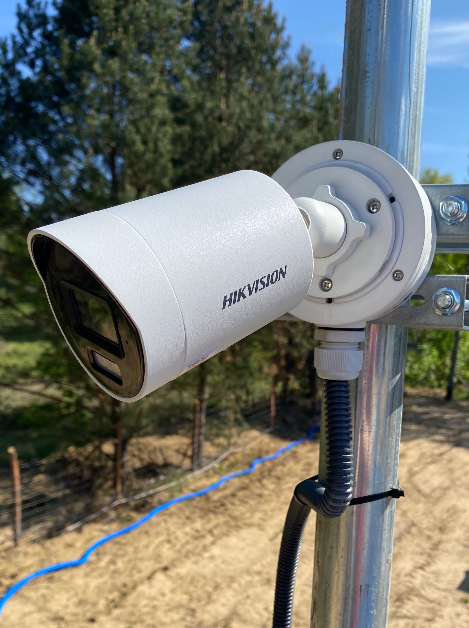 Kamera monitoringu Hikvision zamontowana na metalowym słupku, widoczne przewody w peszlu. W tle drzewa i niebo. Ujęcie z dołu, podkreślające solidność instalacji.
