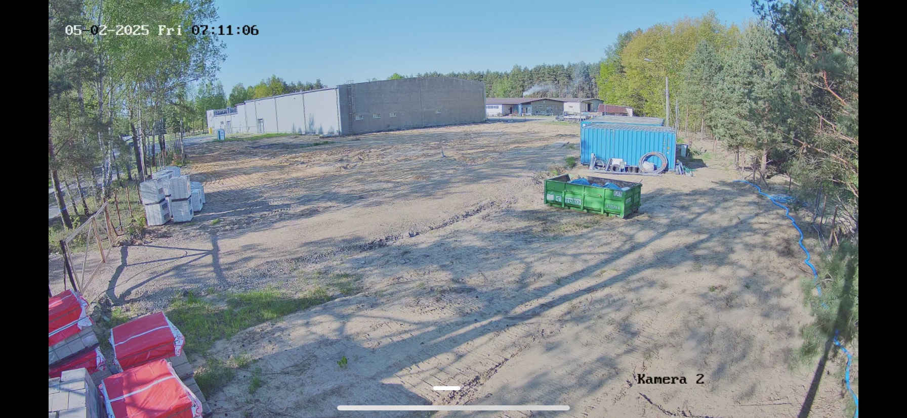 Szeroki kadr z kamery monitoringu: teren budowy z materiałami, kontenerem i budynkiem w tle, oświetlony słonecznym światłem.