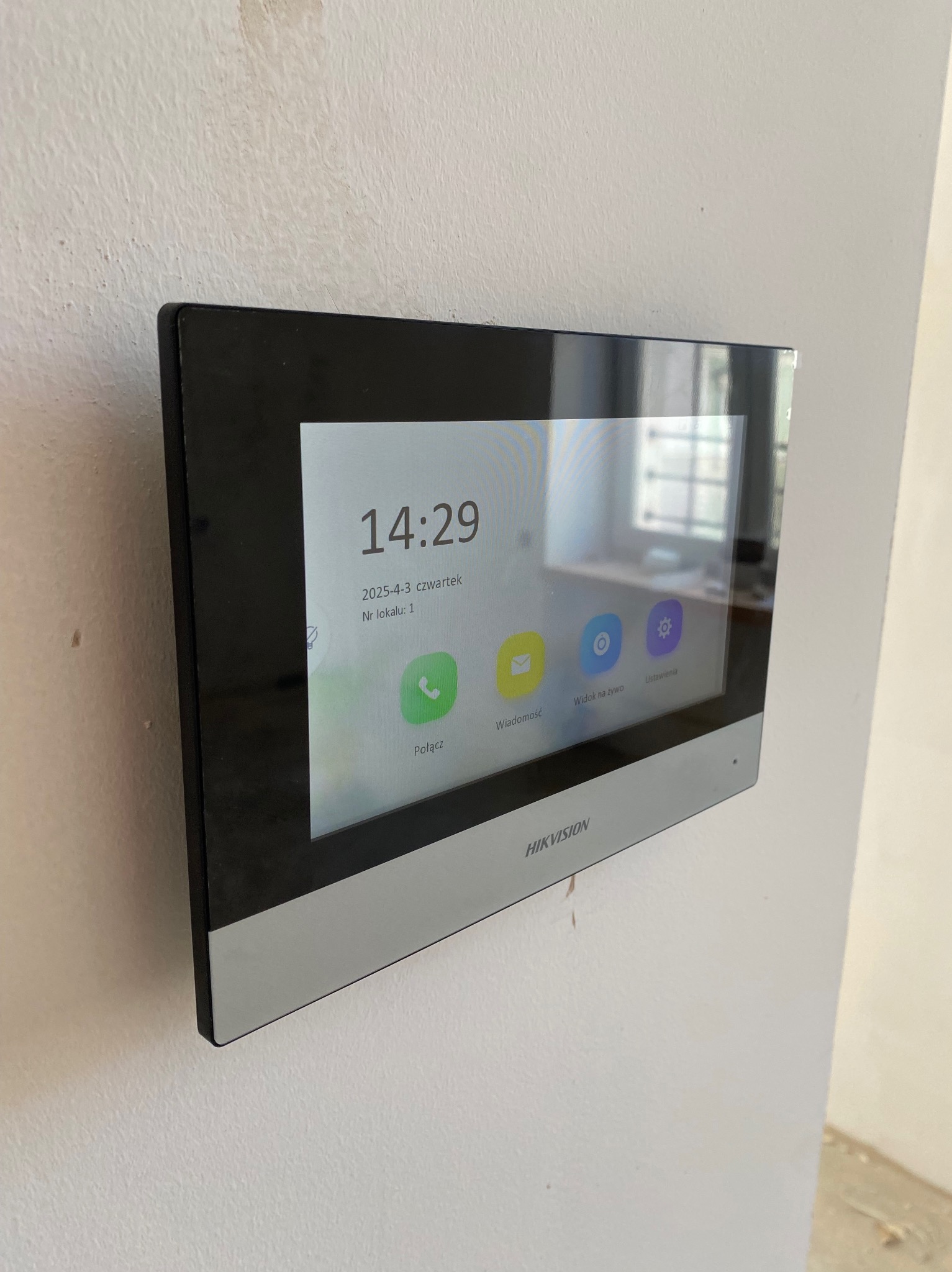 Zamontowany wideodomofon Hikvision na białej ścianie, widoczny ekran z datą i godziną, ikony połączeń i ustawień. Minimalistyczny design, nowoczesny wygląd.