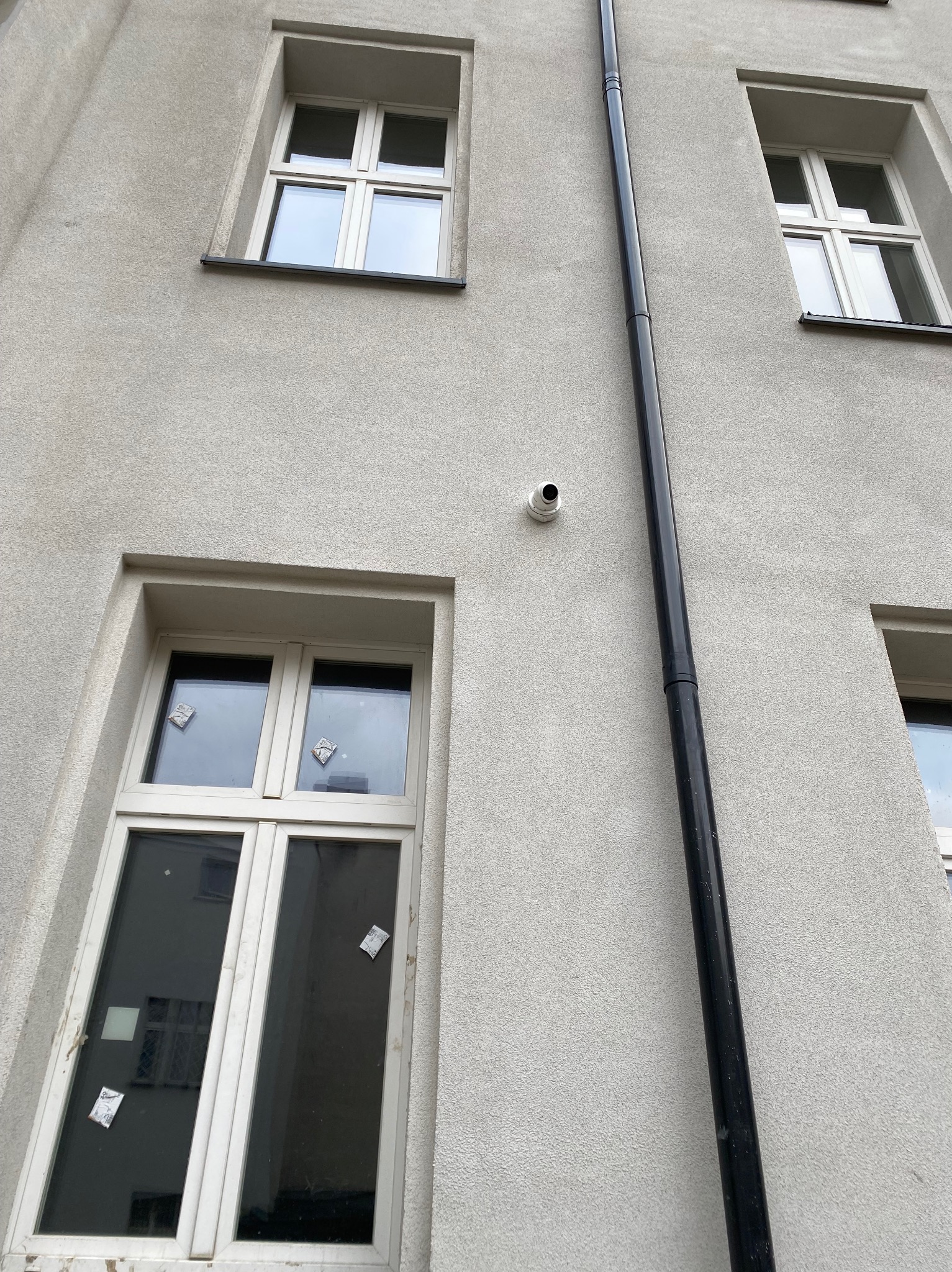 Kamera monitoringu na elewacji budynku z oknami i rurą spustową. Ujęcie z dołu, widoczne okna na różnych kondygnacjach. Szara elewacja.