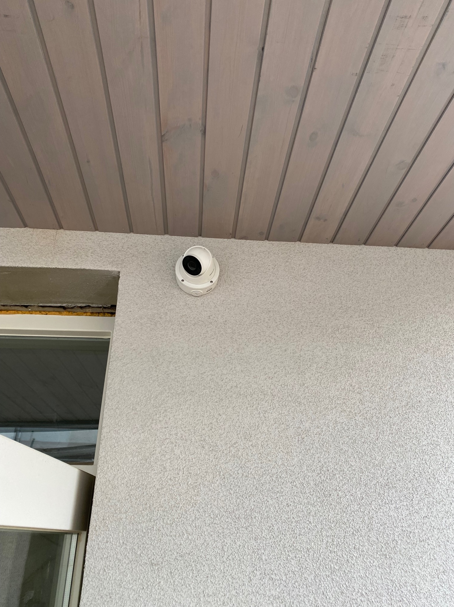 Biała kamera monitoringu kopułkowa zamontowana na jasnej, teksturowanej ścianie pod drewnianym sufitem. Widoczny fragment okna.
