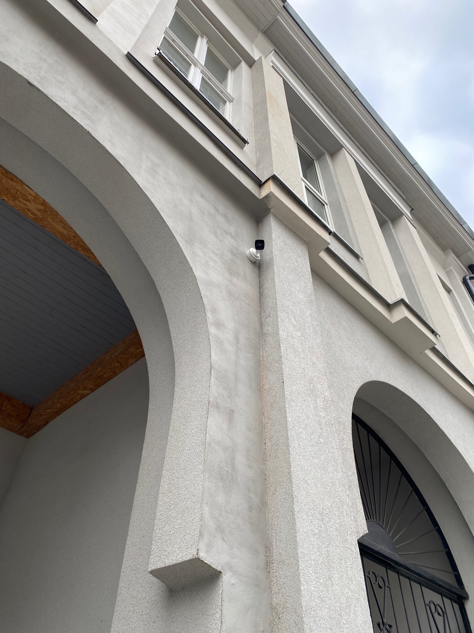 Kamera monitoringu zamontowana na elewacji budynku z łukowymi oknami i szarą strukturą tynku. Widok z dołu, pochmurne niebo w tle.