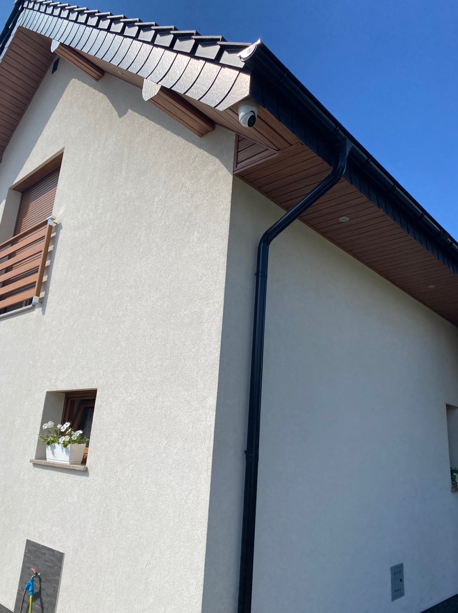 Kamera monitoringu na elewacji domu z szarym dachem i czarną rynną, widoczna z dołu na tle błękitnego nieba w słoneczny dzień. Detal architektoniczny.