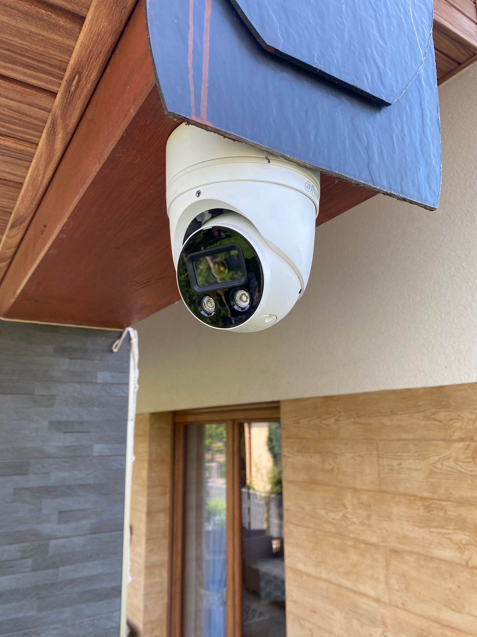 Biała kamera monitoringu Dahua zamontowana pod drewnianym okapem, widok z dołu, z fragmentem elewacji budynku w tle. Widoczne elementy mocujące.