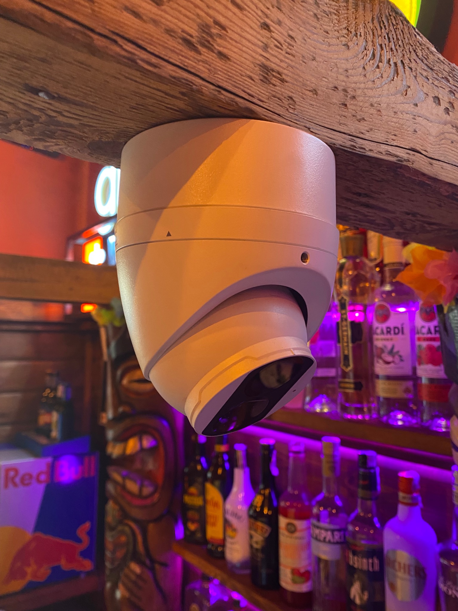 Kamera monitoringu kopułkowa, zamontowana na drewnianej belce w barze, z widocznymi butelkami alkoholu w tle, oświetlonymi fioletowym światłem.