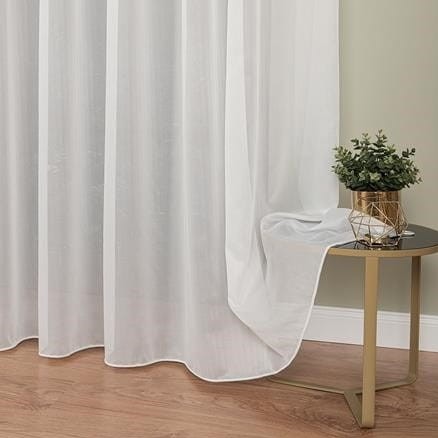 Delikatna, biała firana z subtelnym wzorem, zwiewnie opadająca obok stolika z rośliną w złotym wazonie. Jasne drewno podłogi i stonowana kolorystyka tła.