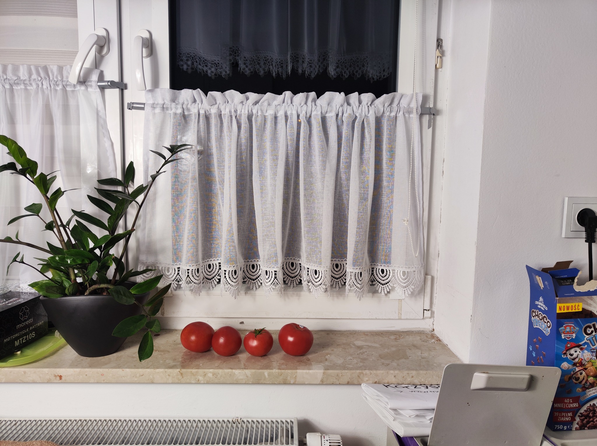 Biała, krótka firanka z koronką w oknie, na parapecie doniczka z kwiatem, cztery pomidory i pudełko płatków śniadaniowych. Ujęcie z bliska, domowe wnętrze.