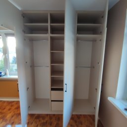 Bartek Wojtasik - Otwarta, biała szafa wnękowa z półkami, drążkami i szufladami, ukazująca wnętrze. Widoczne brązowe panele podłogowe i fragment okna z parapetem.