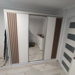 Bartek Wojtasik - Biała szafa przesuwna z drewnianymi panelami w Sępólnie Krajeńskim. Lustrzane drzwi odbijają komodę. Widoczna podłoga z paneli i białe drzwi.