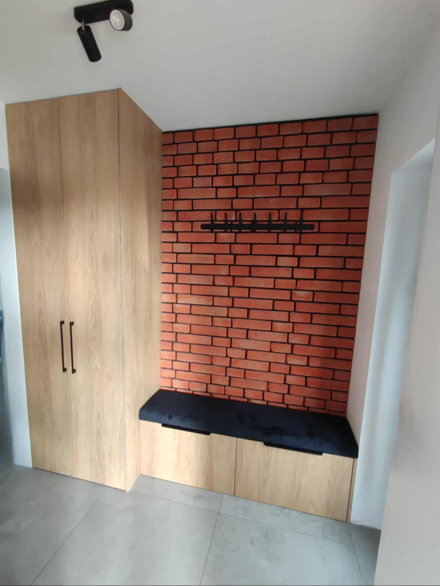 Funkcjonalna szafa z siedziskiem i wieszakiem na tle ściany z cegły w przedpokoju. Jasne drewno, czarne detale, nowoczesny design.