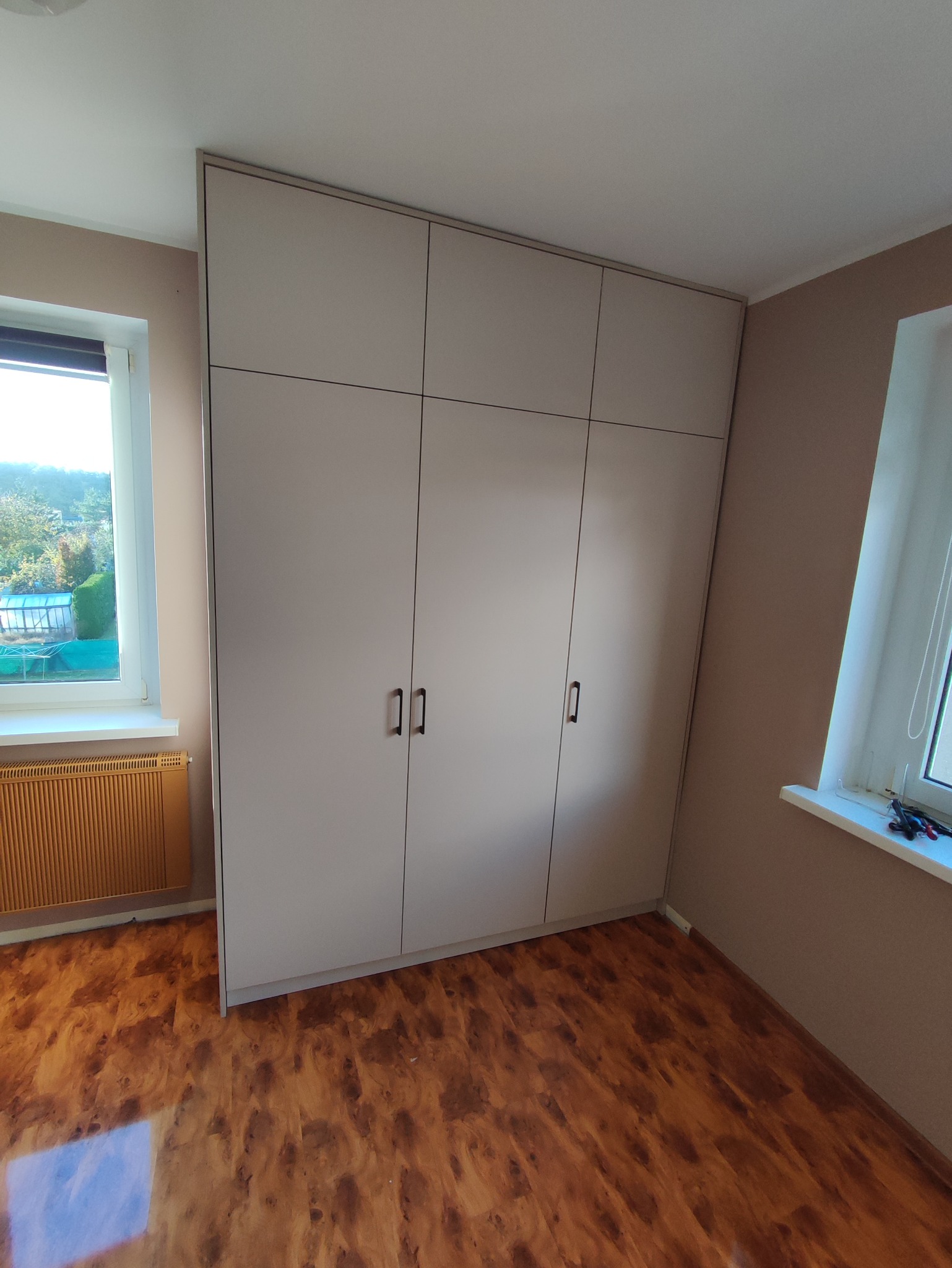 Jasnoszara szafa wnękowa z trzema segmentami, każdy z dwoma drzwiami, w pokoju z drewnianą podłogą i oknami. Minimalistyczny design, proste uchwyty.