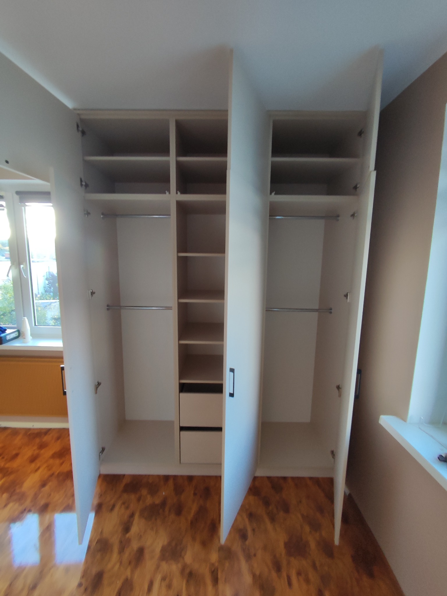 Otwarta, biała szafa wnękowa z półkami, drążkami i szufladami, ukazująca wnętrze. Widoczne brązowe panele podłogowe i fragment okna z parapetem.