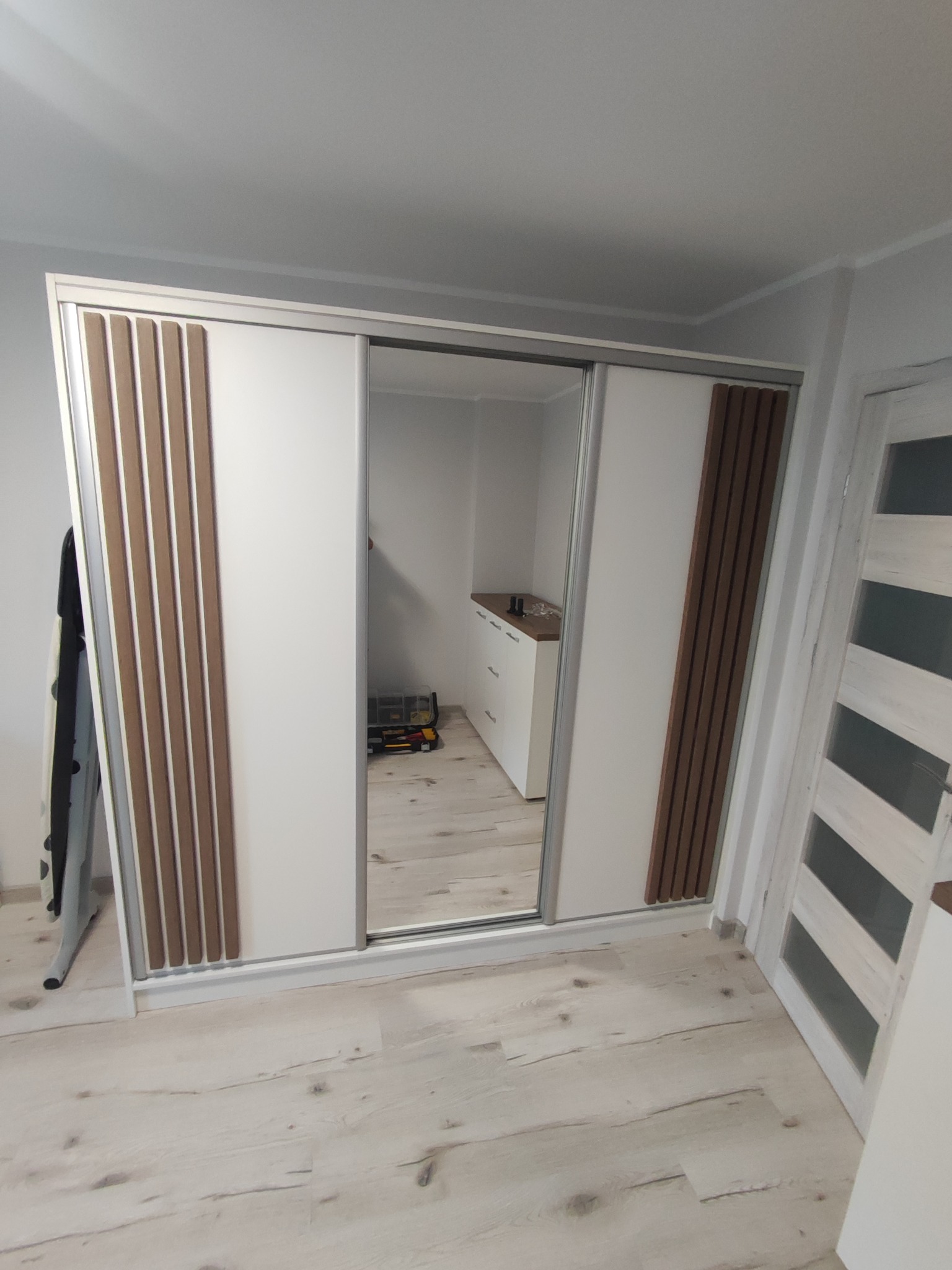 Biała szafa przesuwna z drewnianymi panelami w Sępólnie Krajeńskim. Lustrzane drzwi odbijają komodę. Widoczna podłoga z paneli i białe drzwi.