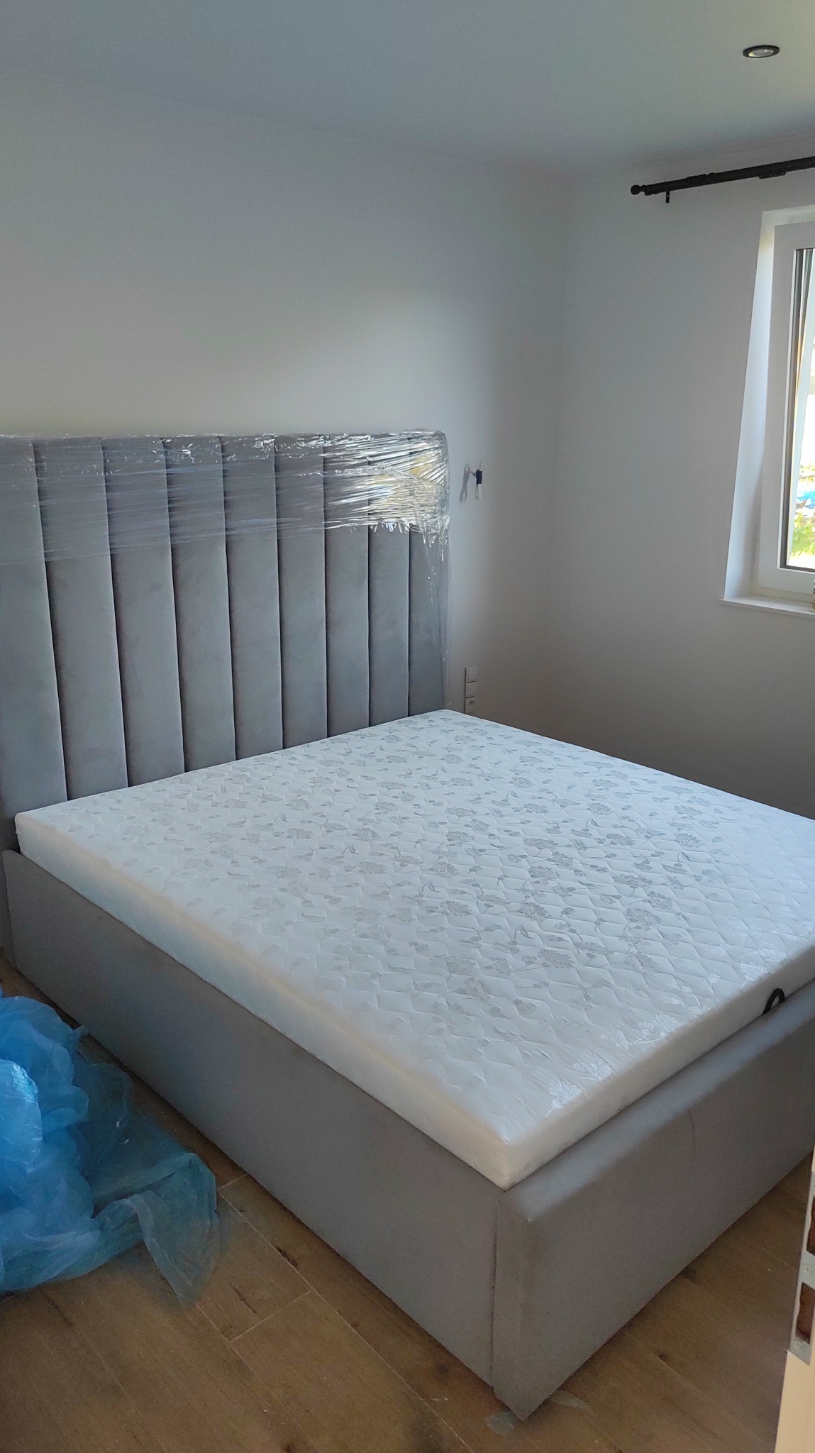 Szare łóżko tapicerowane z wysokim wezgłowiem w folii ochronnej, materac z wzorem, jasne wnętrze pokoju, widok z boku, drewniana podłoga, okno z zasłoną.