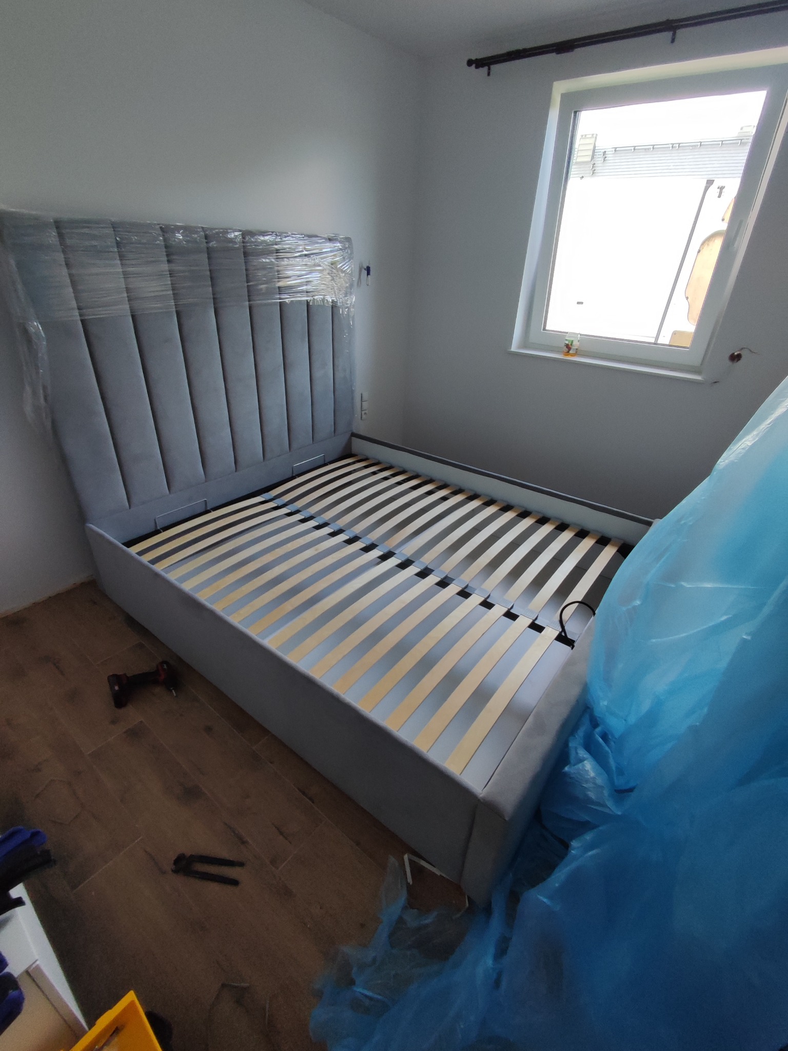 Szare, tapicerowane łóżko z wysokim wezgłowiem w folii ochronnej, z drewnianym stelażem, w trakcie montażu w jasnym pokoju z oknem. Widoczne narzędzia.