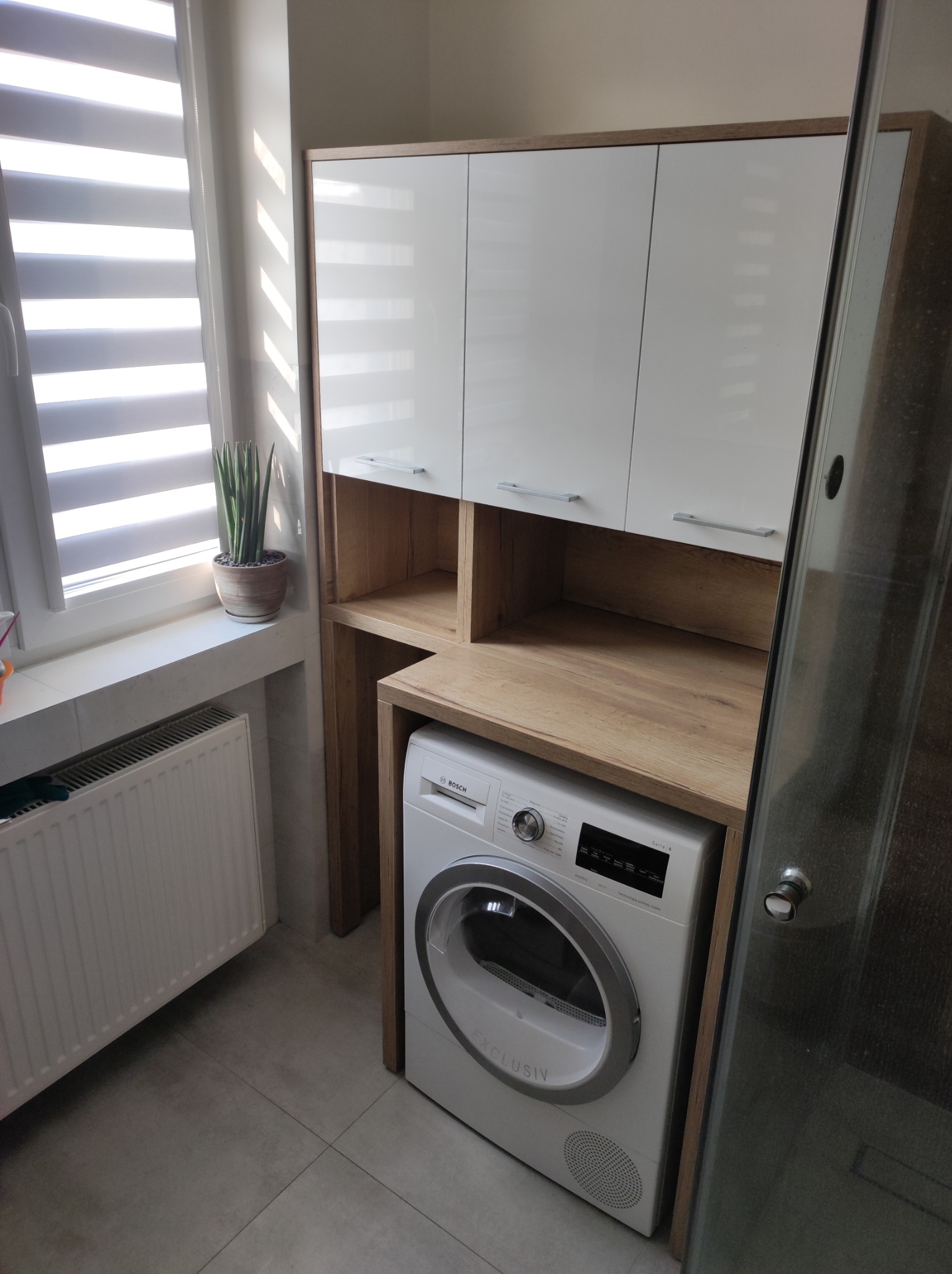 Białe szafki wiszące z drewnianymi półkami nad pralką Bosch, tworzące zabudowę w łazience. Widoczny fragment okna z żaluzjami i grzejnik.
