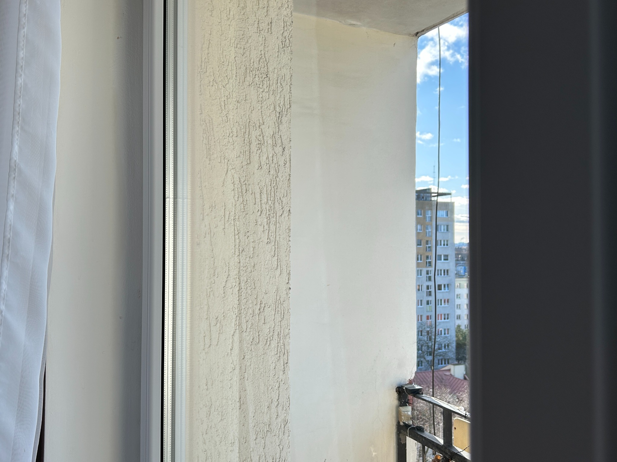 Widok z balkonu przez czyste okno na budynek mieszkalny w słoneczny dzień. Biała firanka po lewej stronie kadru. Fragment balustrady balkonu.