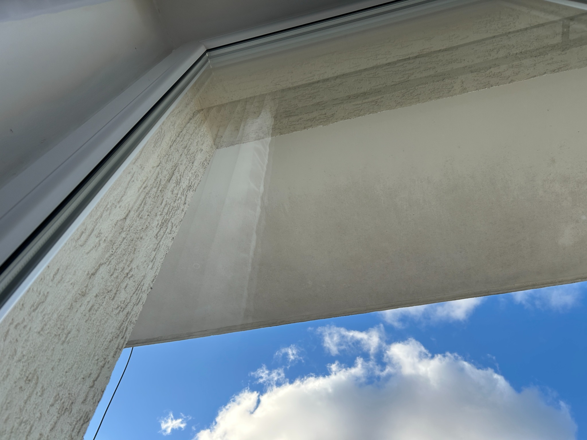 Brudne okno z widokiem na błękitne niebo i chmury, uchwycone pod kątem. Widoczny fragment ramy okiennej i ściany z fakturą.