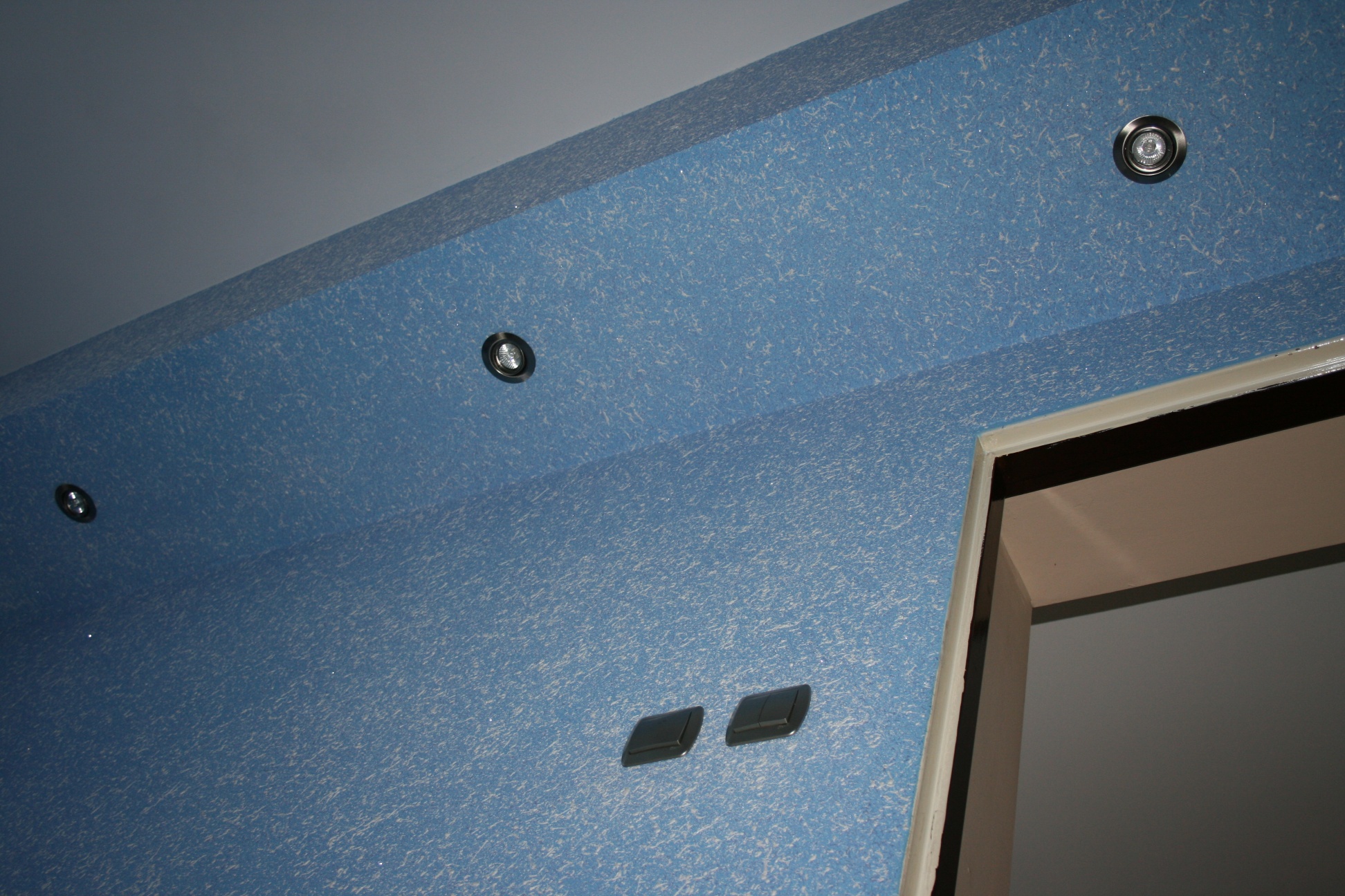 Niebieska ściana i sufit pokryte tapetą natryskową z widocznymi punktami oświetleniowymi i włącznikami światła, fragment futryny drzwiowej w prawym dolnym rogu.