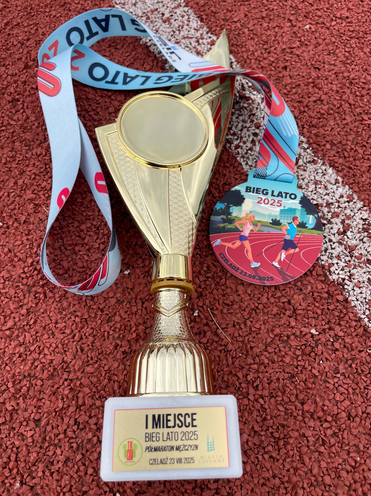 Złoty puchar i medal z napisem Bieg Lato 2025 leżą na czerwonej bieżni. Trofeum za pierwsze miejsce w półmaratonie męskim, Czeladź.