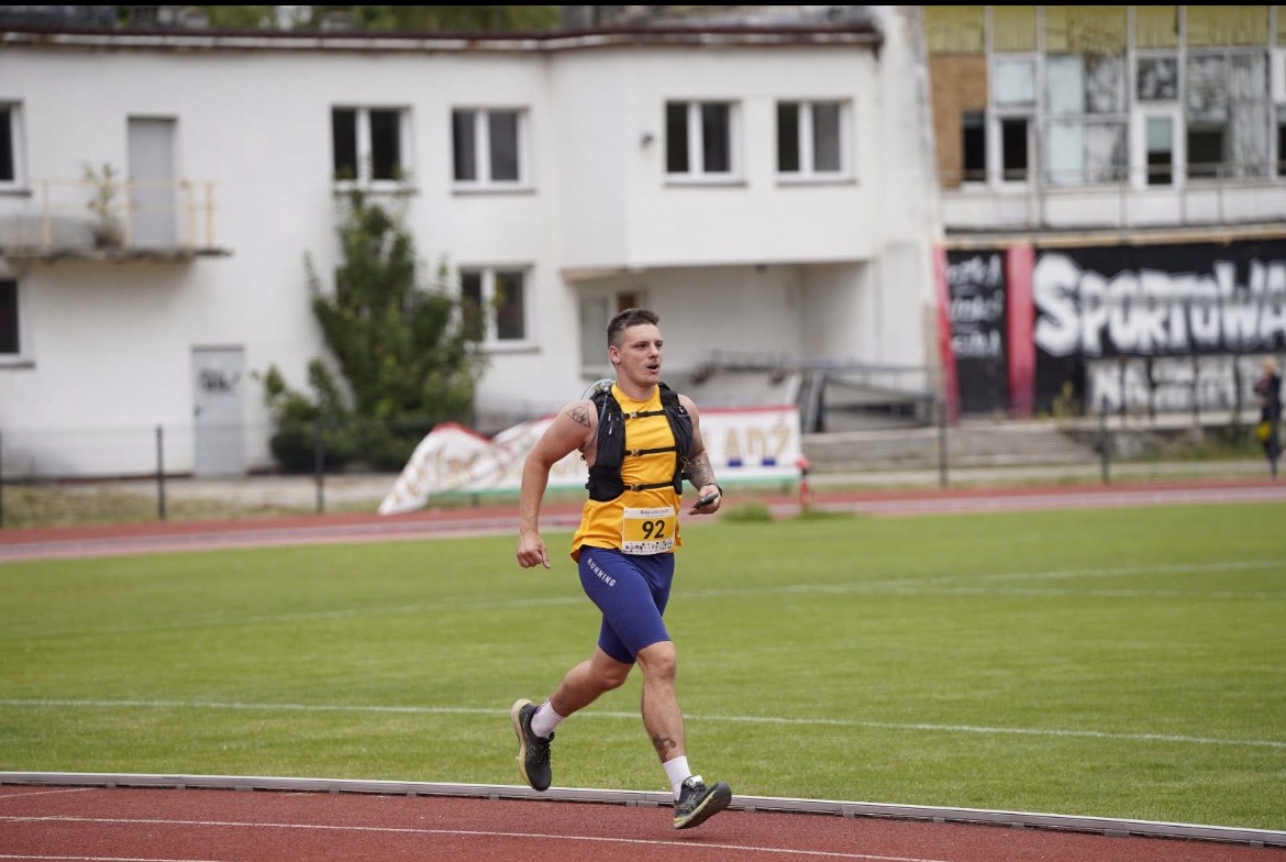 Mężczyzna w żółtej koszulce i niebieskich spodenkach biegnie po bieżni stadionu. Widoczny numer startowy 92 i budynek w tle. Dynamiczne ujęcie w trakcie treningu biegowego.