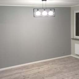 HeiderStudio - Szare, świeżo pomalowane wnętrze pokoju z drewnopodobną podłogą, białym grzejnikiem pod oknem i nowoczesną lampą sufitową. Minimalistyczny design.