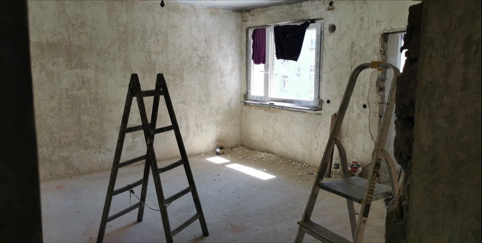 Pokój w trakcie remontu: ściany po szpachlowaniu, dwie drabiny, okno z suszącymi się ubraniami. Widoczne ślady prac remontowych na podłodze i ścianach.