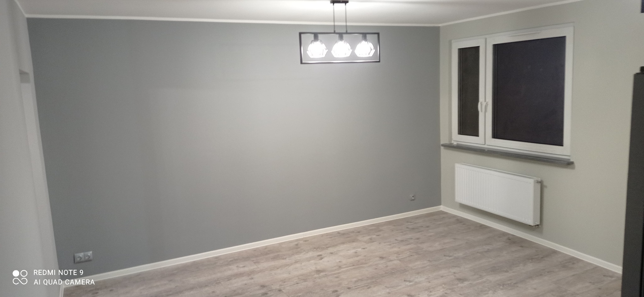 Szare, świeżo pomalowane wnętrze pokoju z drewnopodobną podłogą, białym grzejnikiem pod oknem i nowoczesną lampą sufitową. Minimalistyczny design.