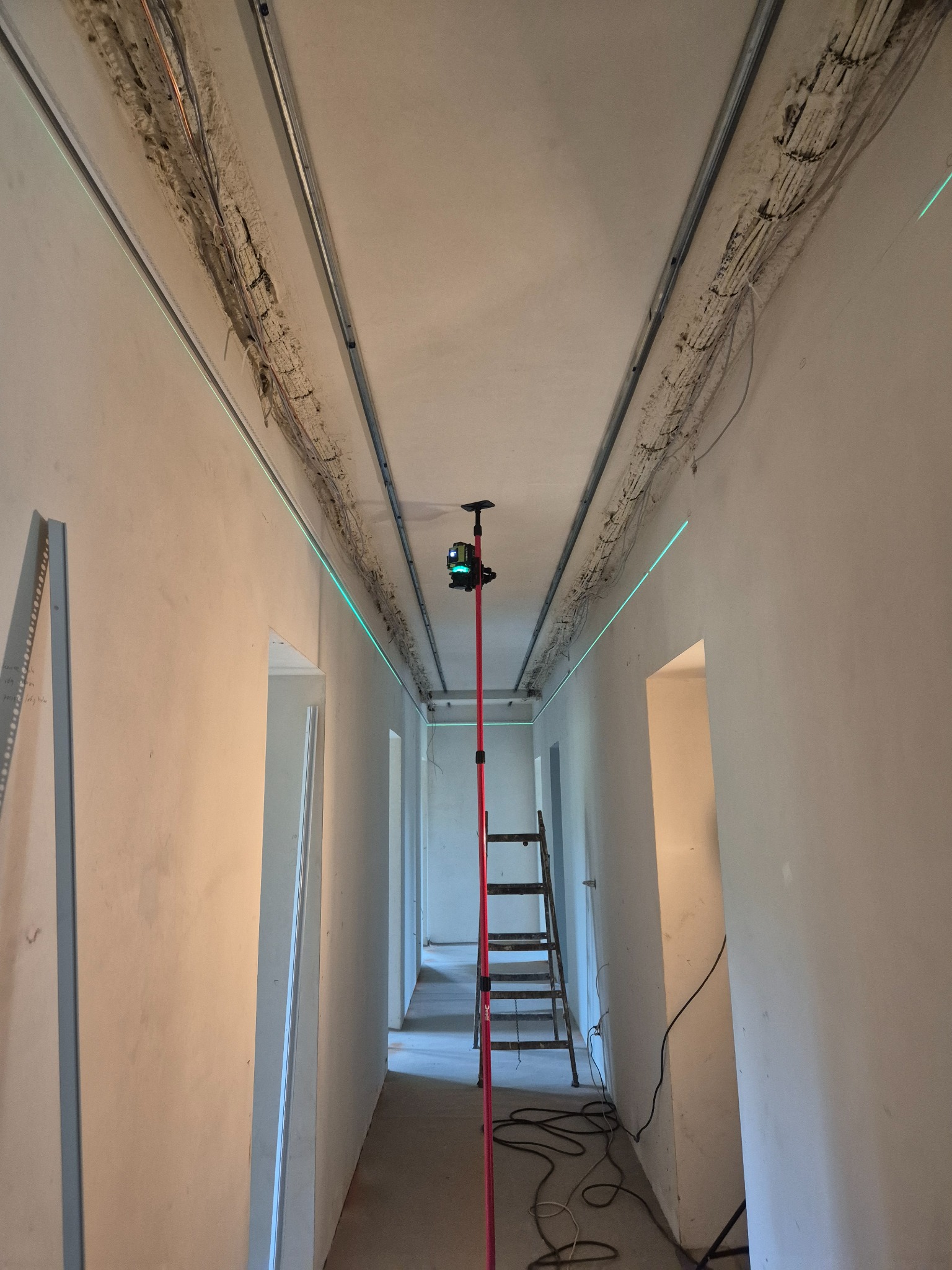 Wnętrze w trakcie prac wykończeniowych: widoczne profile sufitowe, instalacje elektryczne i poziomica laserowa wyznaczająca linię. Ściany przygotowane do dalszych prac.