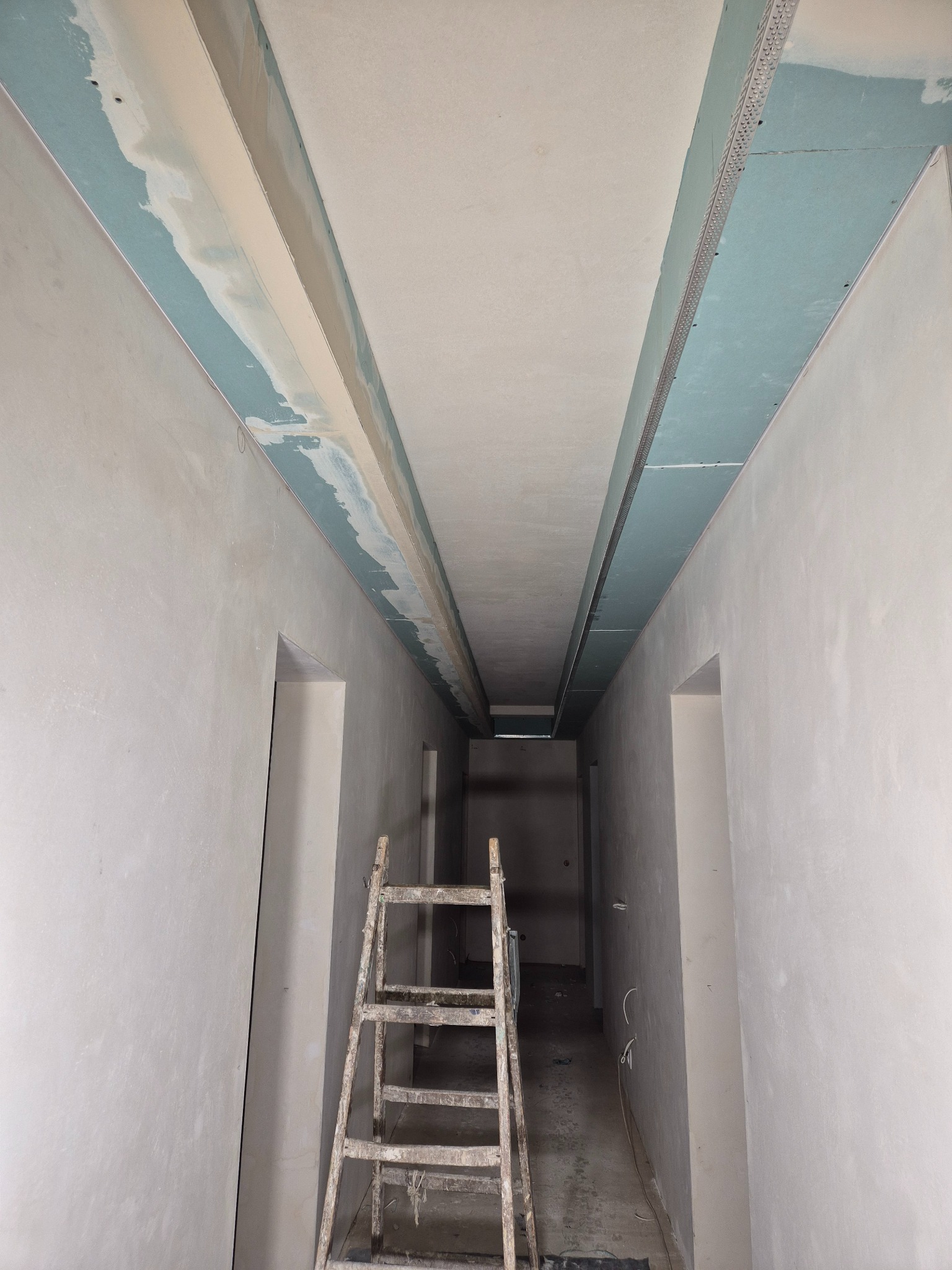 Korytarz w trakcie prac wykończeniowych: widoczne płyty gipsowo-kartonowe na suficie, ściany przygotowane do szpachlowania, drabina w centralnej części kadru.