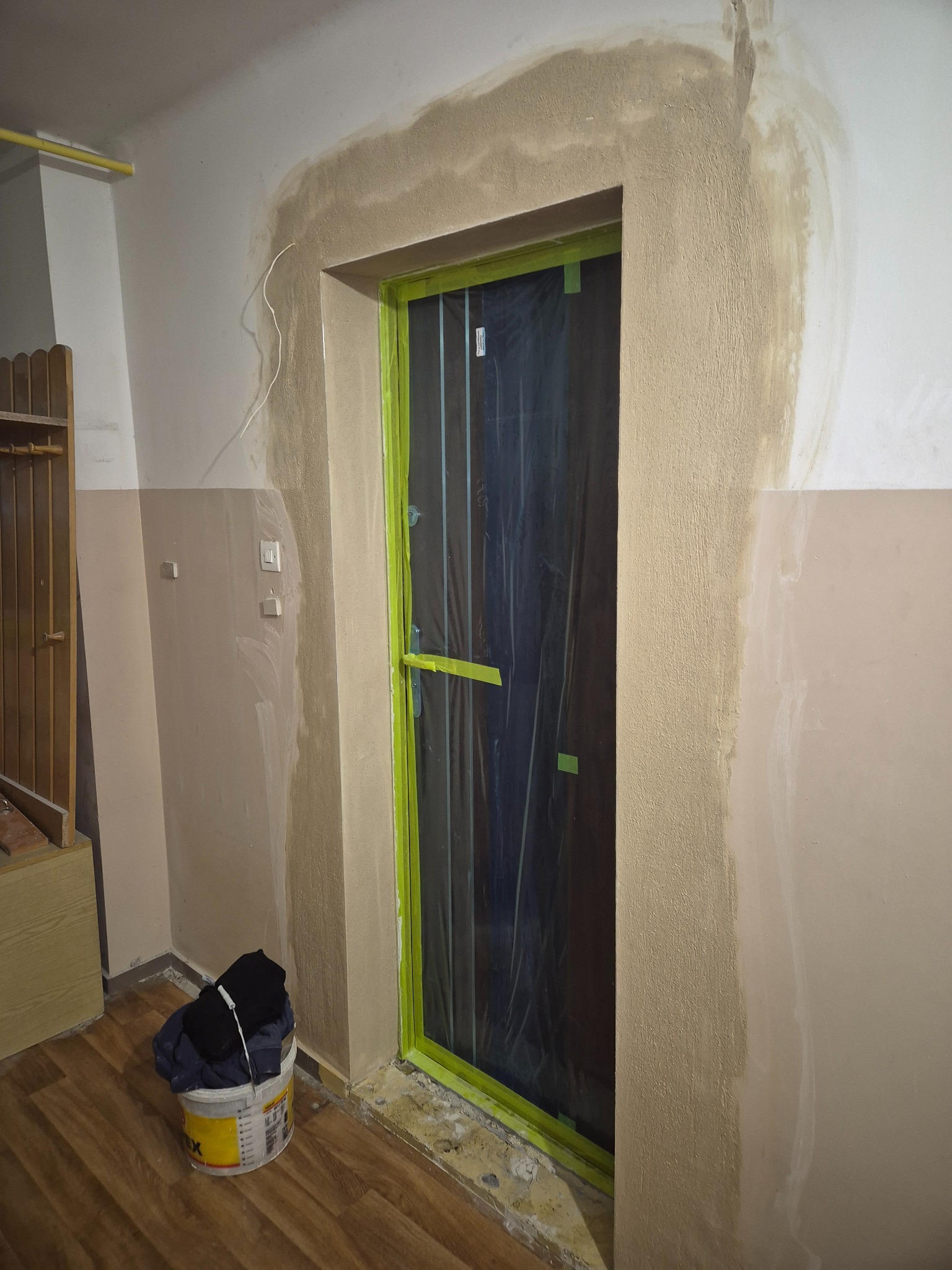 Przygotowanie do malowania: obramowanie drzwi po szpachlowaniu, zabezpieczone folią i taśmą malarską. Widoczne ślady szpachli na ścianie i wiadro z farbą.